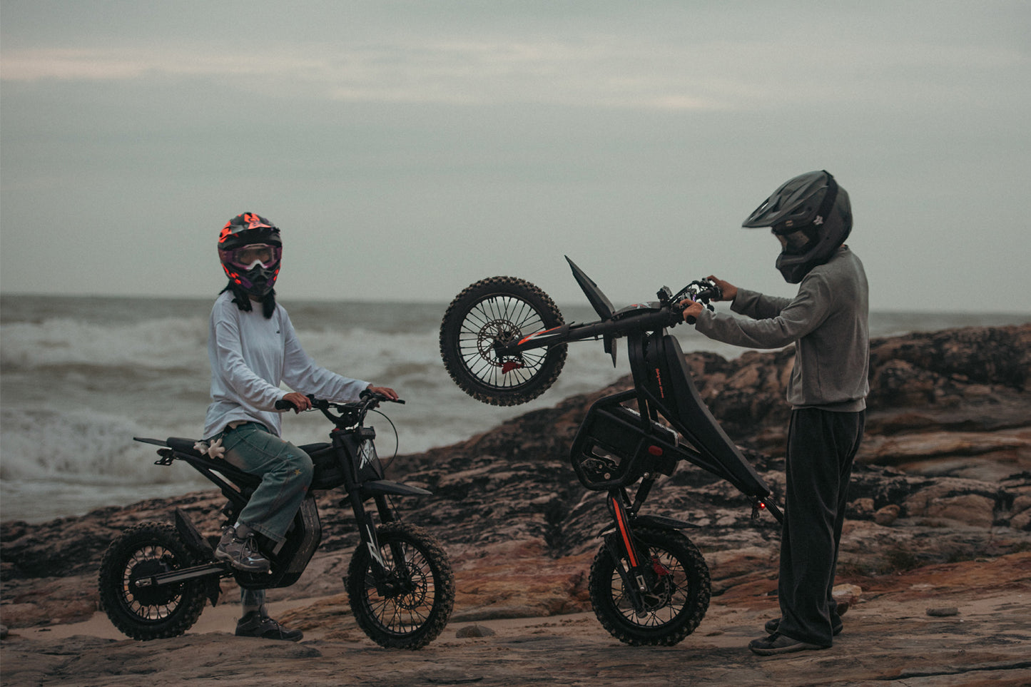 Two people with dirt bikes on a rocky beach with ocean waves in the background
